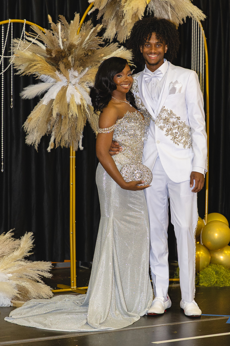 Couple posing at a formal event: woman in a silver glitter gown with a round clutch, man in a white suit with gold embroidery, gold decor in the background.