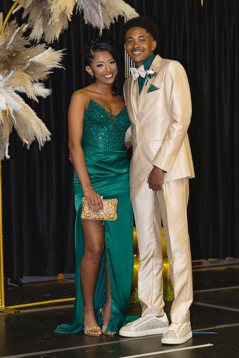 Couple in formal wear posing together: woman in emerald green gown with a gold clutch, man in a beige suit and matching sneakers.