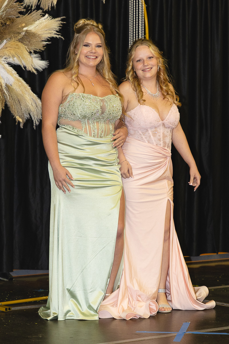 Two women in pastel formal dresses pose and smile on a stage with a black curtain backdrop and decorative plants nearby.