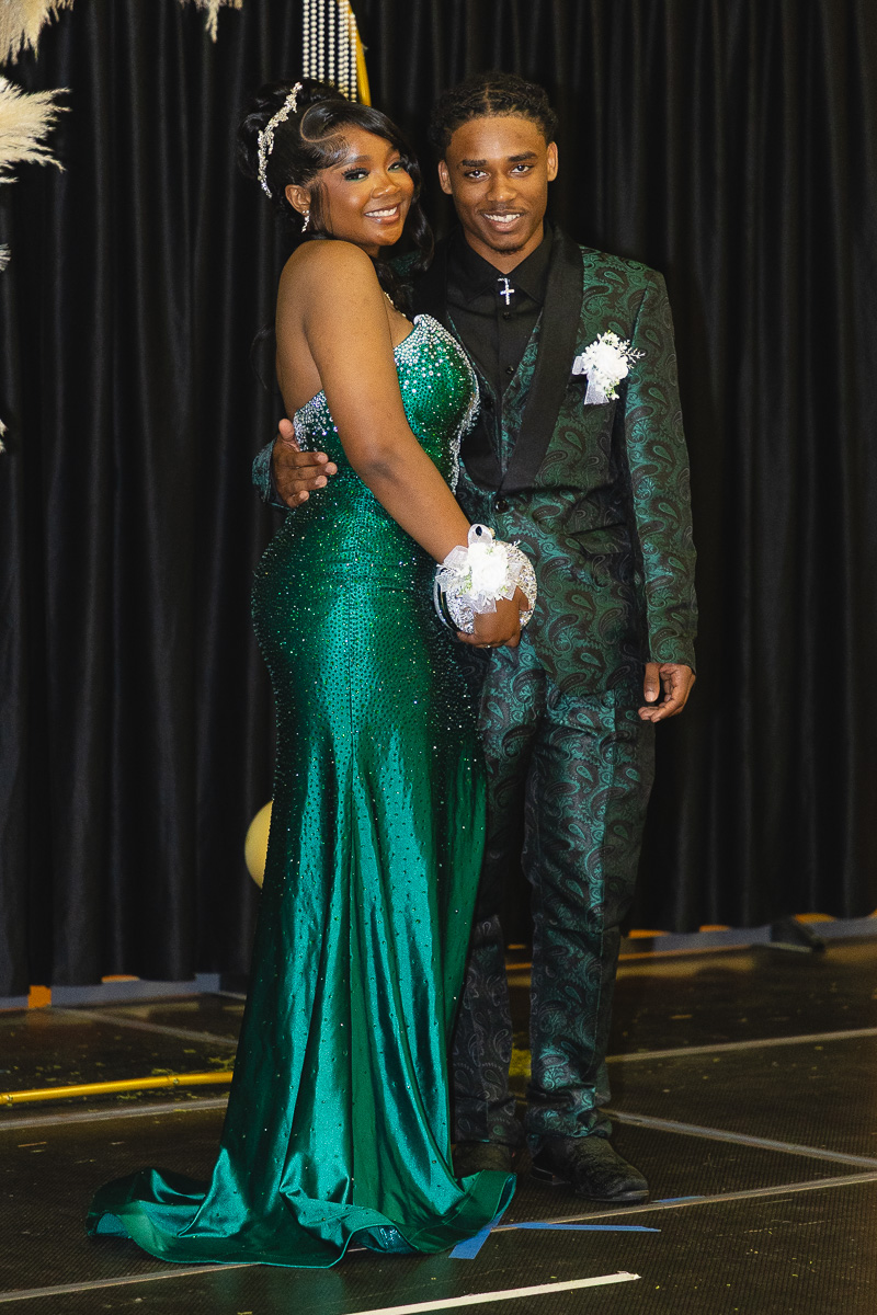 Smiling couple posing on a stage in formal attire: a woman in an emerald green sequined gown with a corsage and a man in a green patterned suit, both with arms around each other.