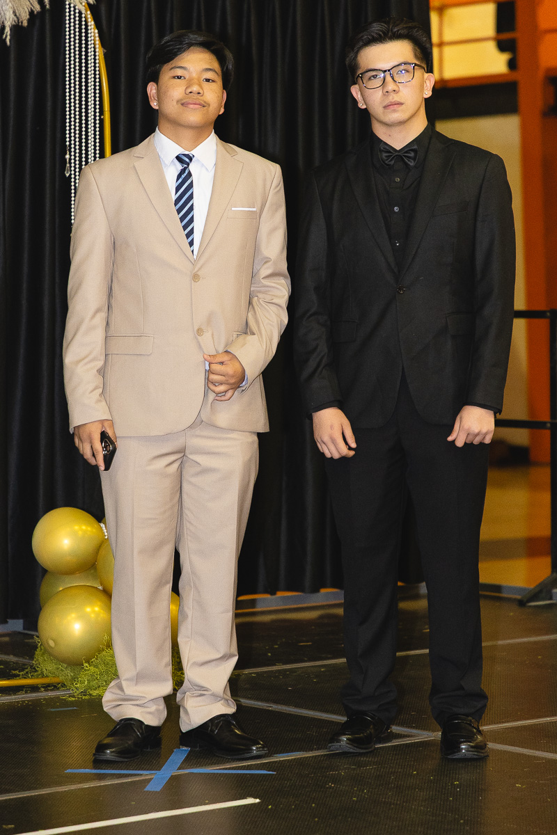 Two young men in formal suits pose on a stage, one in a beige suit and the other in a black suit, with gold balloons nearby on the left.