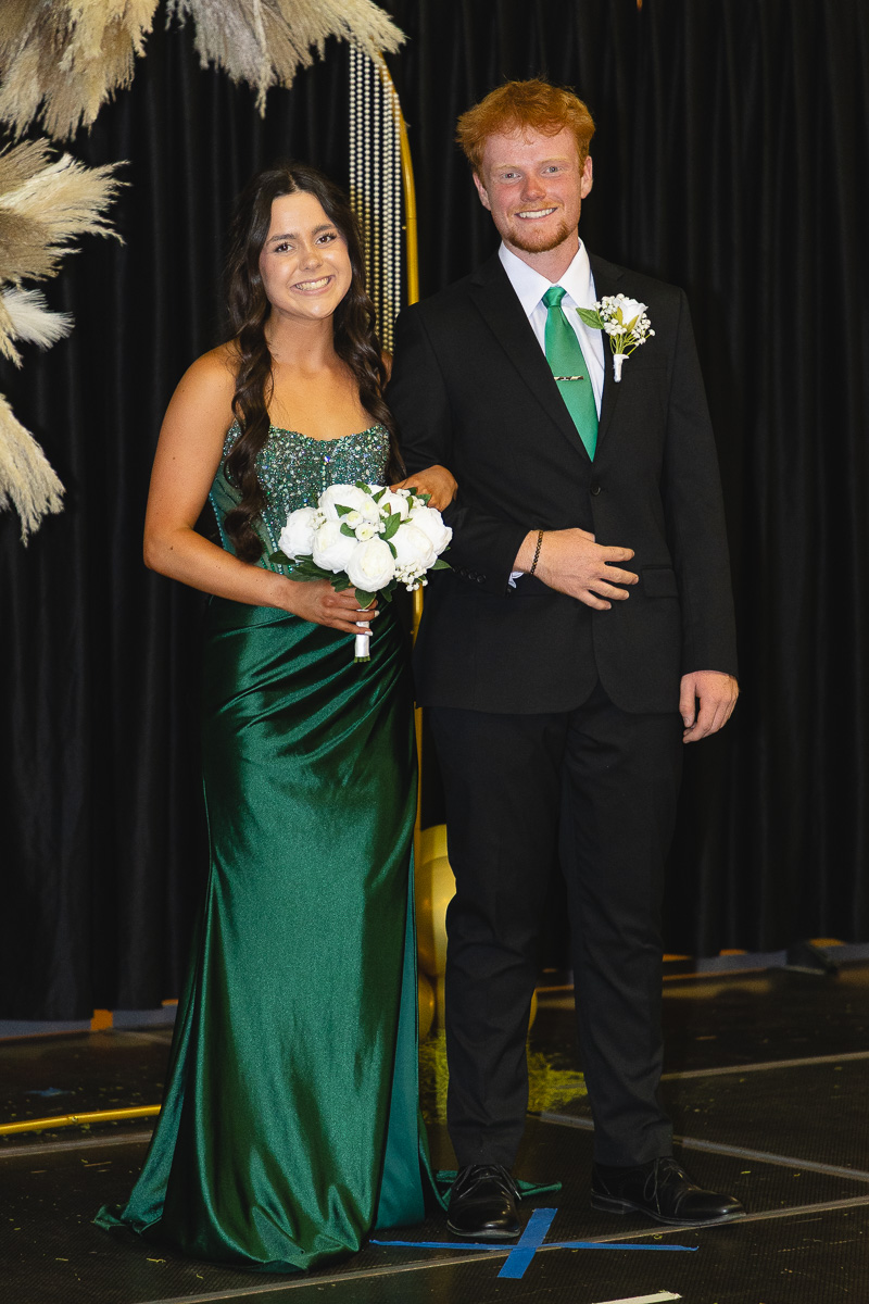 Couple in formal attire posing on stage, the woman in a green gown holding a white bouquet and the man in a black suit with a green tie and boutonniere.