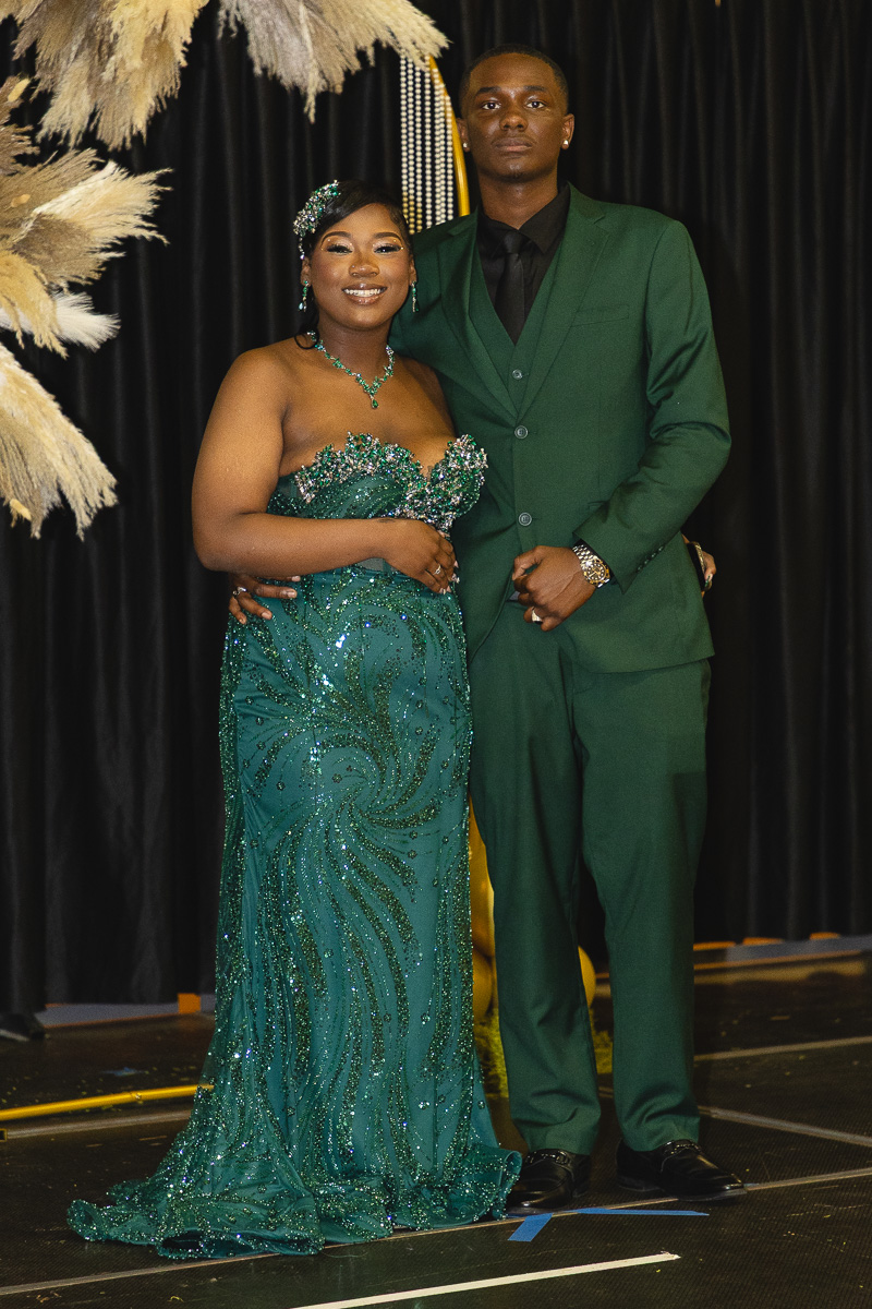 Couple in formal emerald attire posing on a stage with decorative pampas grass backdrop.