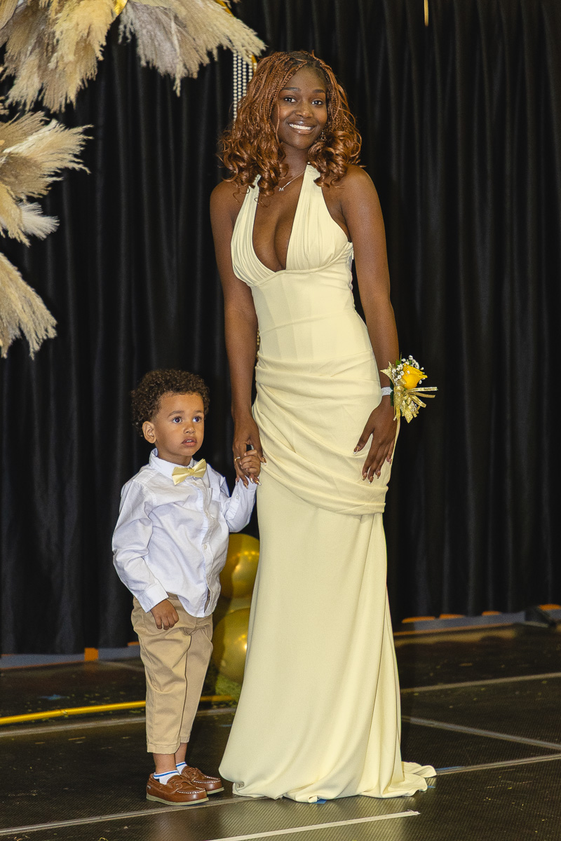 Smiling woman in a pale yellow gown stands on a stage, holding hands with a young boy in a white shirt and beige pants beside her on a black-curtained backdrop.