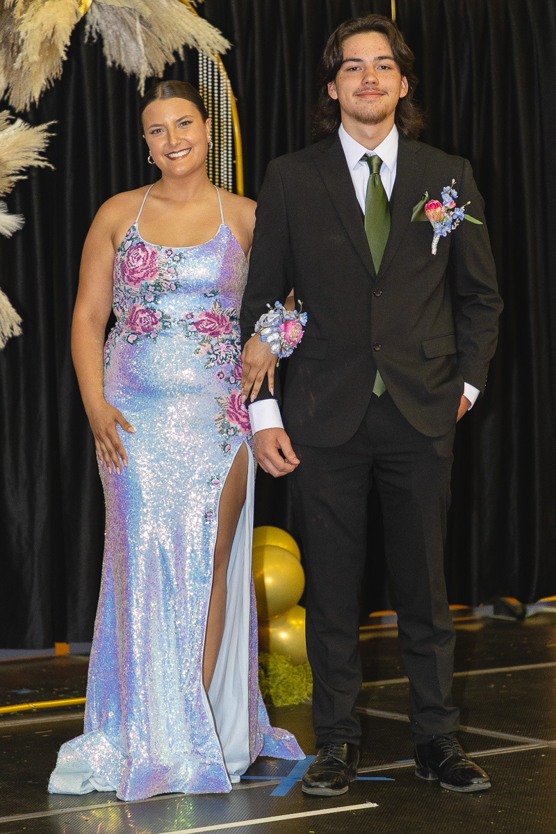 Two young people in formal attire pose arm-in-arm on a stage, smiling after prom or formal event. The woman wears a sparkly lavender dress; the man wears a black suit with a green tie and boutonniere.