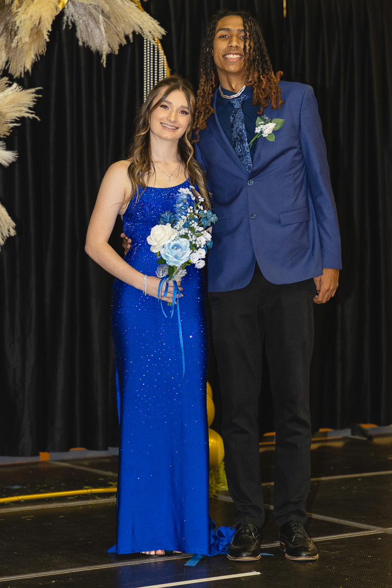 Prom couple posing on a stage: girl in a sparkling royal blue gown holding a white-and-blue bouquet, boy in a blue blazer and black pants.