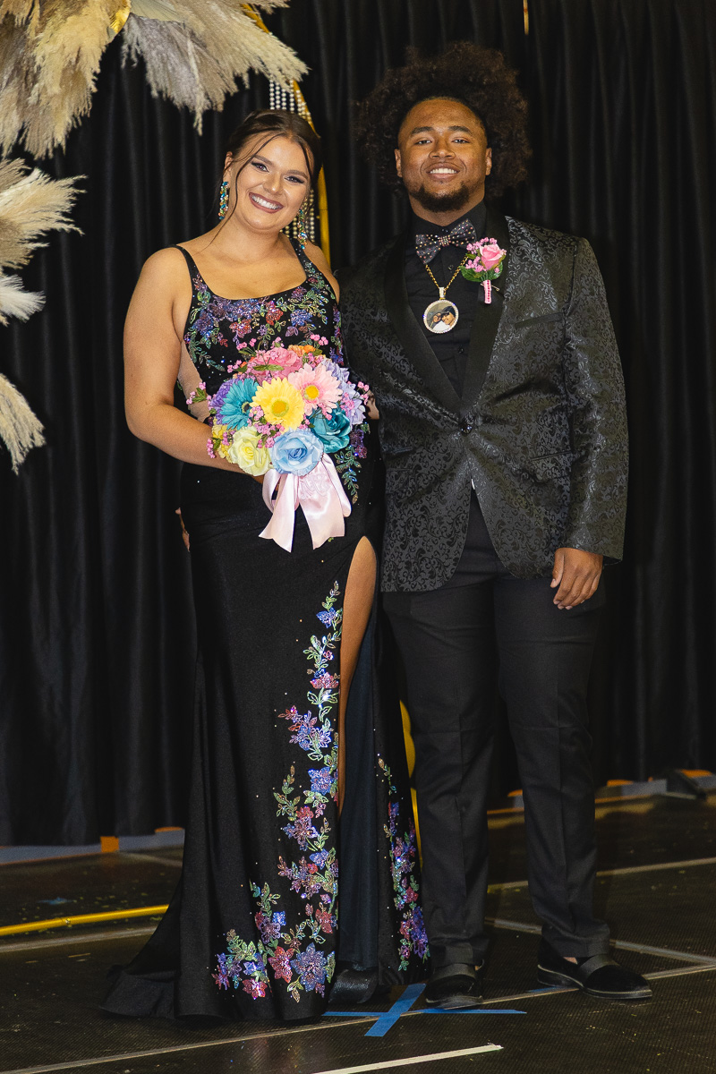 Smiling couple on a stage in formal wear; woman in a floral black gown holds a colorful bouquet, man in a patterned suit beside her.