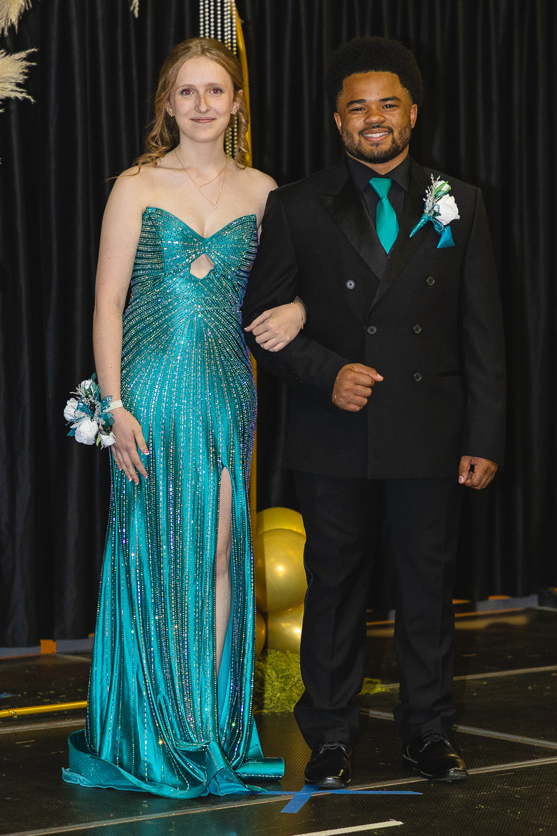 Young woman in a turquoise sequined gown with a thigh-high slit stands arm-in-arm with a man in a black suit and teal tie at a formal event, corsage on her wrist, balloons in the background.