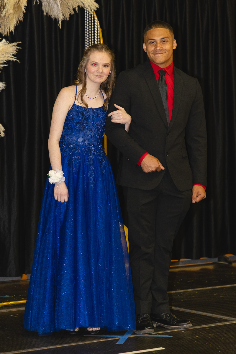 Young couple posing on stage: woman in a blue ball gown with a corsage, man in a black suit with a red shirt, arm linked.