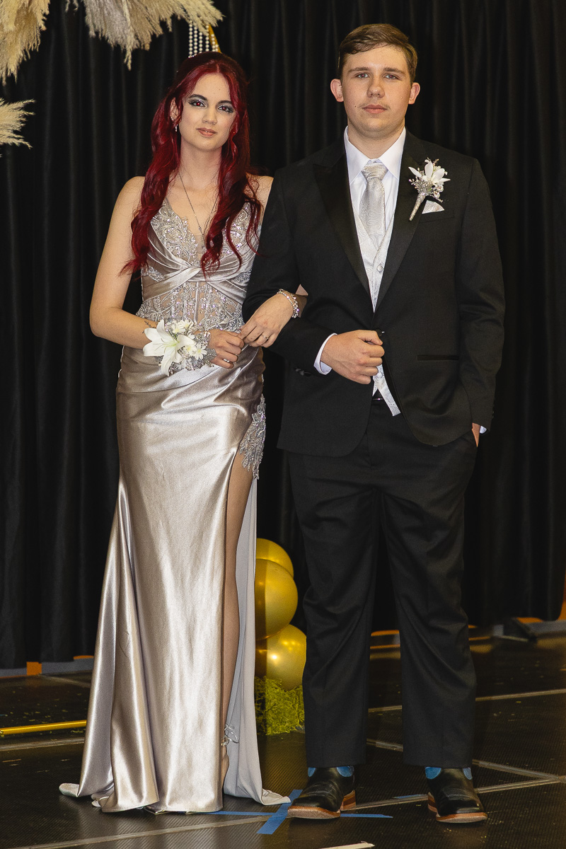 Couple in formal attire pose on a stage, the woman in a silver gown with a thigh-high slit and corsage, and the man in a black suit with a boutonniere, arm-in-arm.