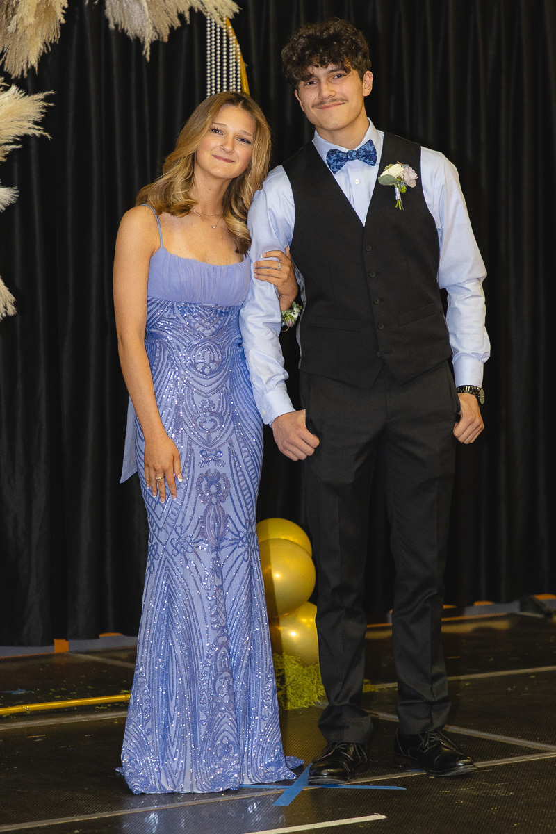 Two young adults posing arm-in-arm at a formal event; woman in a light blue sequined gown and man in a black vest with a blue bow tie and boutonniere.