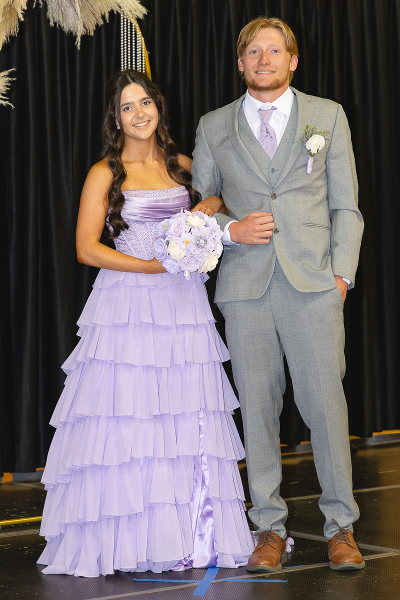 Couple posing on a stage: woman in lavender tiered gown holding a bouquet, man in gray suit with lavender tie and boutonniere, arm in arm against a black curtain backdrop.
