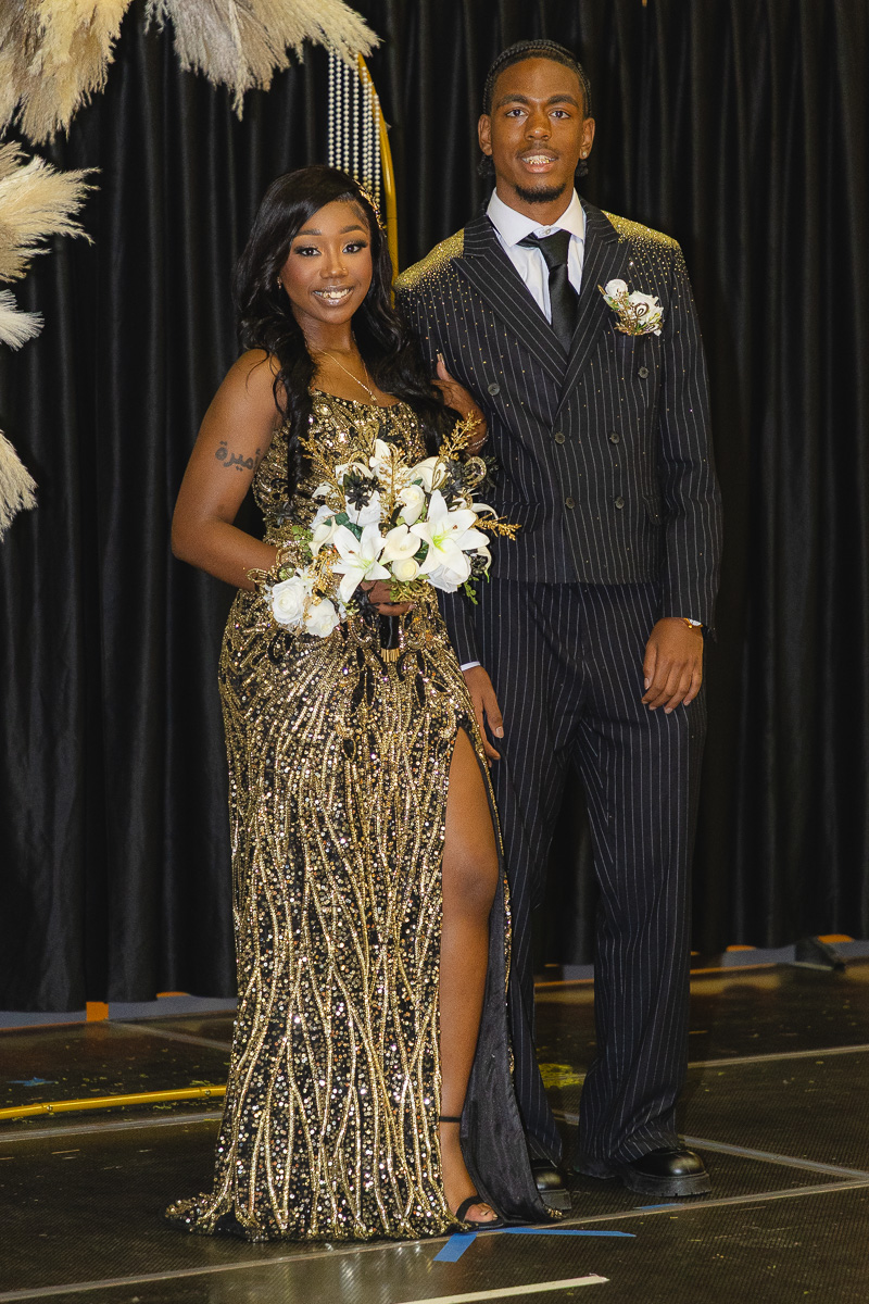 Couple posing on a stage: woman in a gold sequined gown with a thigh-high slit holding a white bouquet, man in a dark pinstripe suit with a boutonniere.