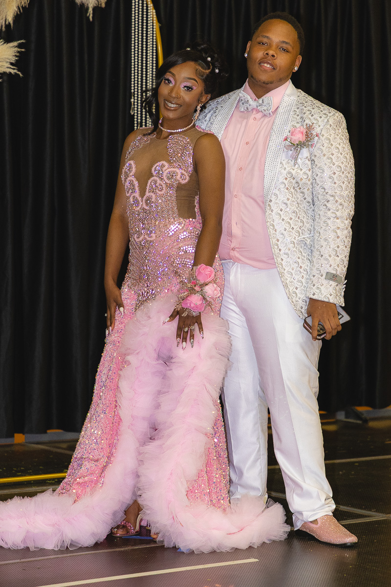 Couple in formal attire posing on stage; woman in a sparkling pink gown with a fluffy pink train and corsage, man in a light patterned blazer and pink shirt.