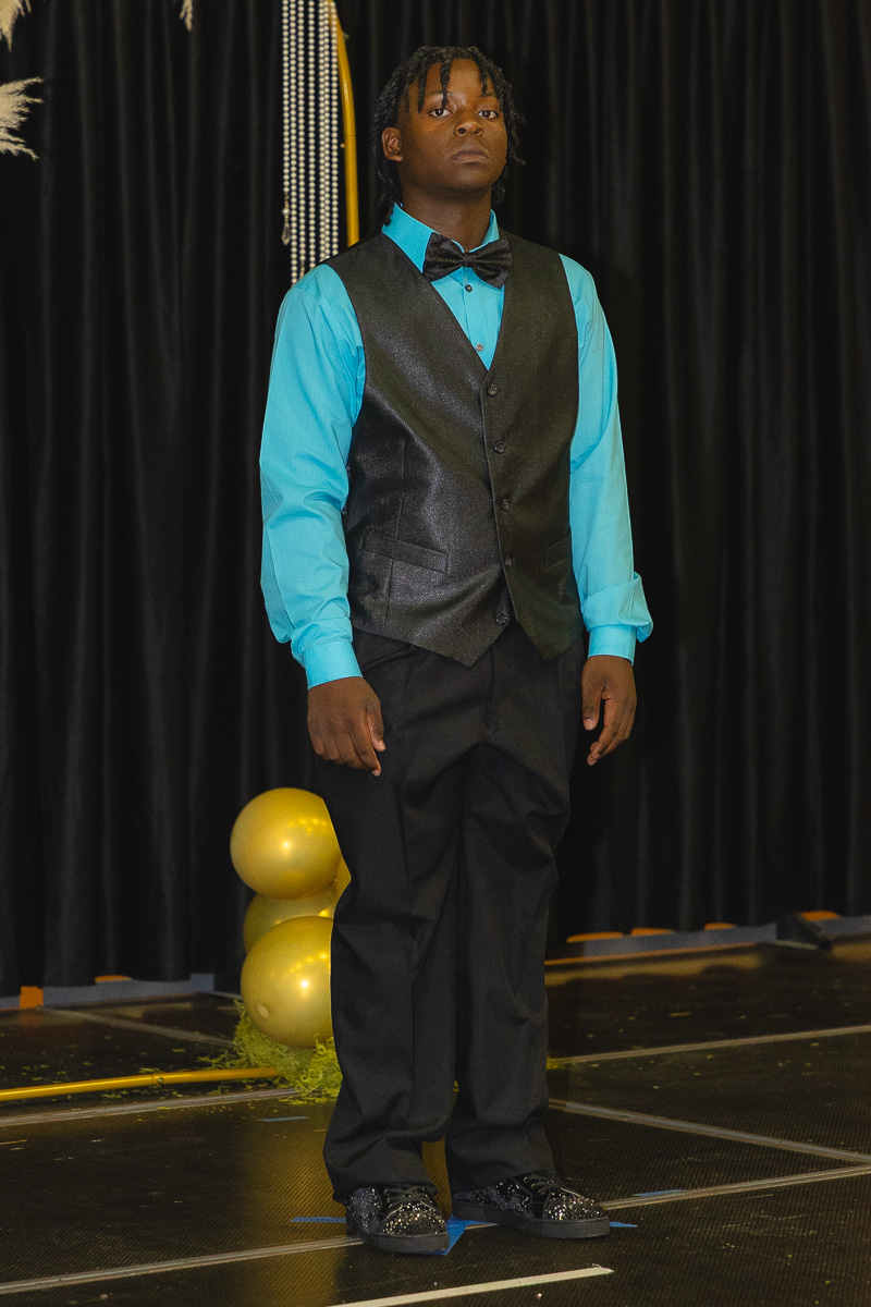 Young man on stage in a teal shirt, black vest and bow tie, facing forward on a dim backdrop with balloons nearby.