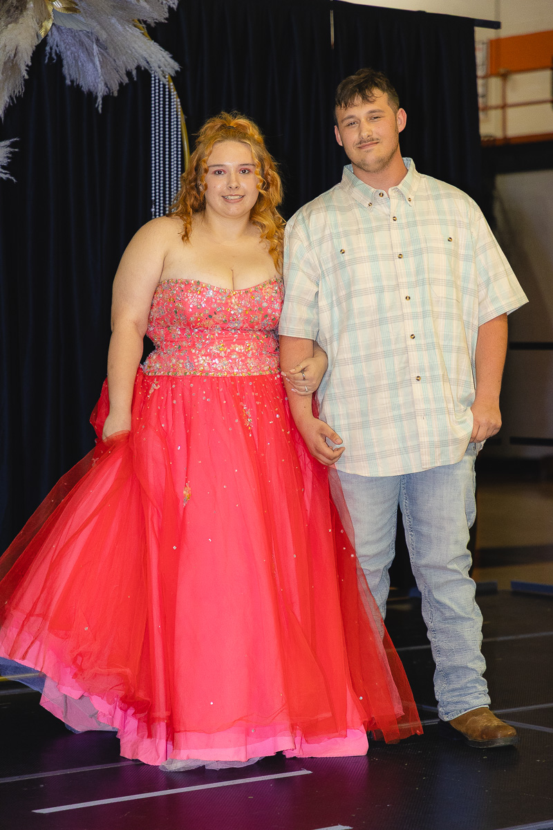 Couple posing on a stage: woman in a bright pink strapless ball gown beside a man in a light plaid shirt and jeans, holding arms outward.