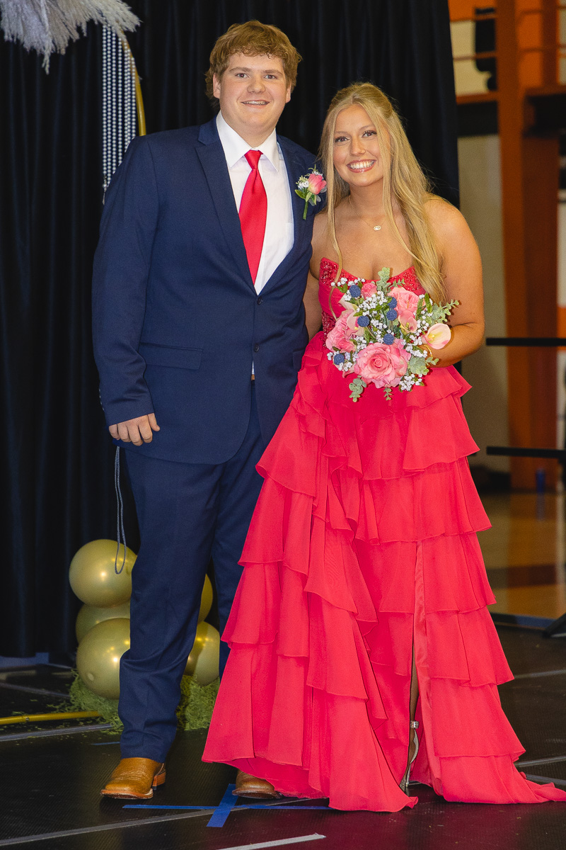 Young couple posing on a stage after a formal event: man in a navy suit with a red tie and boutonniere, woman in a bright pink tiered gown holding a bouquet.