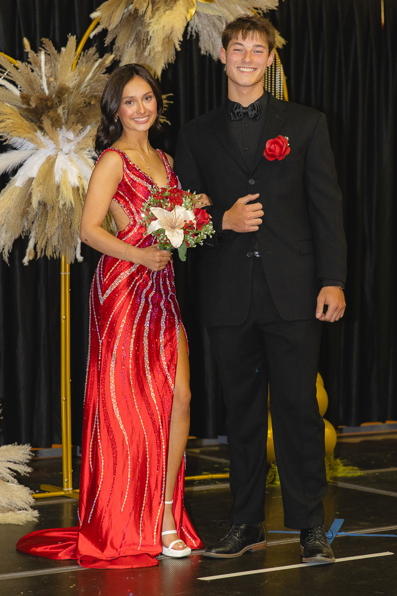 Young couple in formal wear posing on a stage: woman in a red beaded gown holding a bouquet, man in a black tuxedo with a red boutonniere.