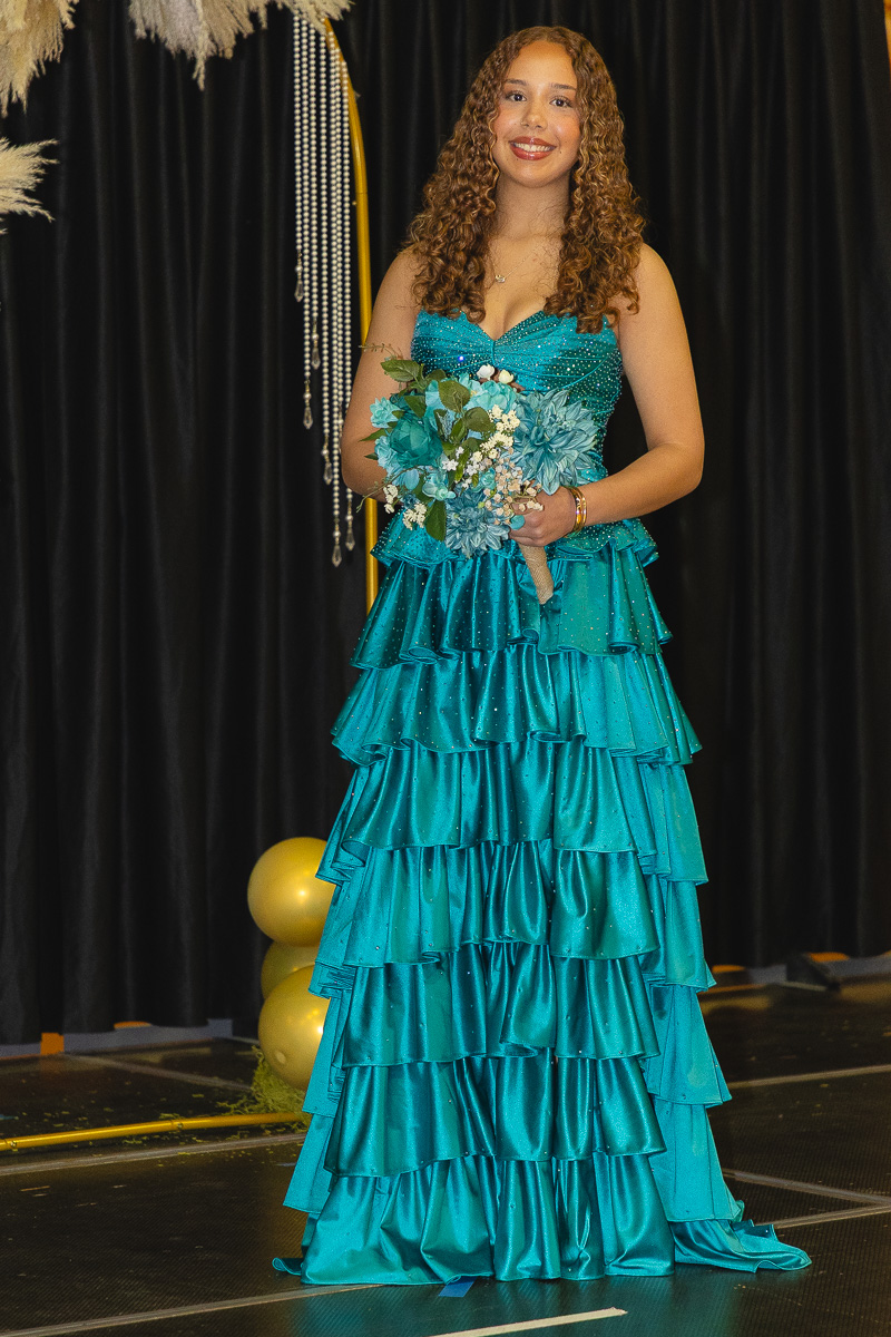 Young woman in a teal tiered gown holding a bouquet of blue flowers on a stage with a black curtain backdrop and gold balloons behind her.