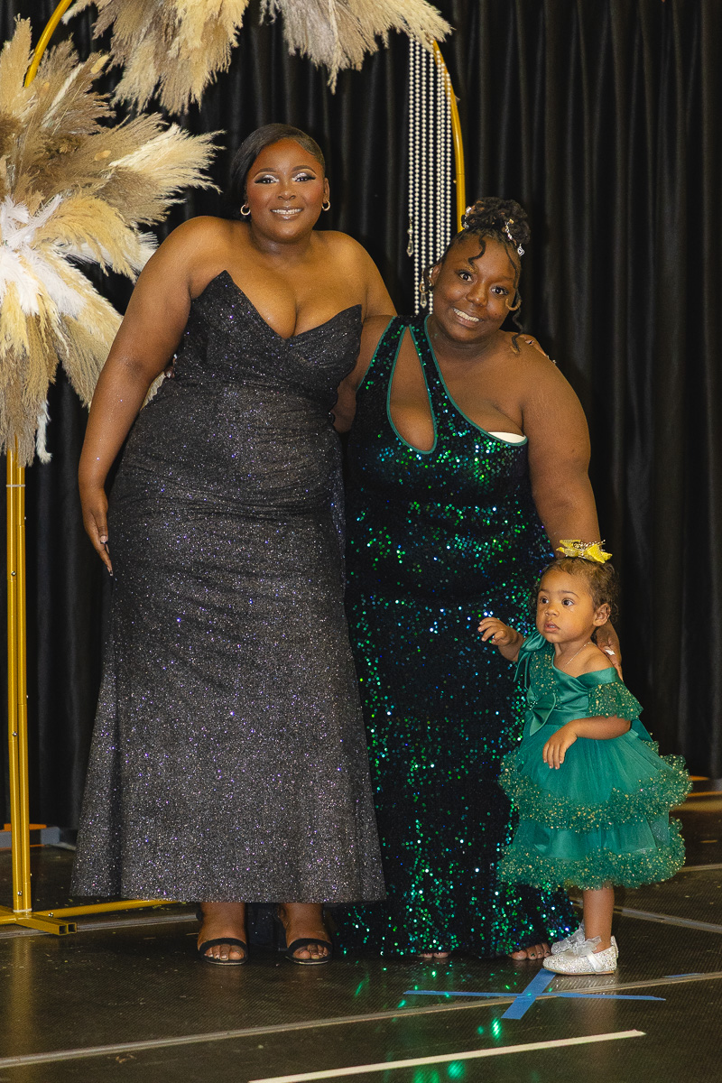 Three women posing on a stage in formal gowns, with a young girl in a green tutu-style dress beside them.