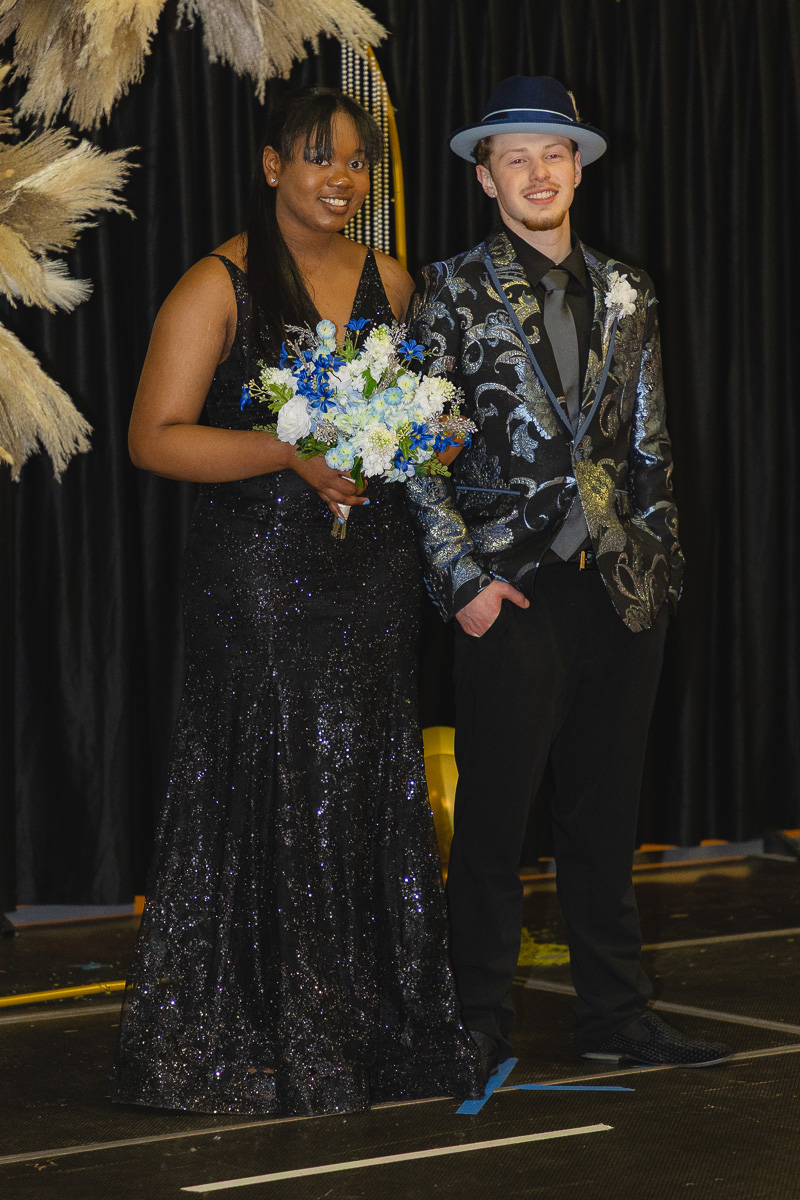 Young couple in formal attire posing on a stage; woman in a black sequined gown holding a blue‑white bouquet, man in a patterned suit and blue hat.