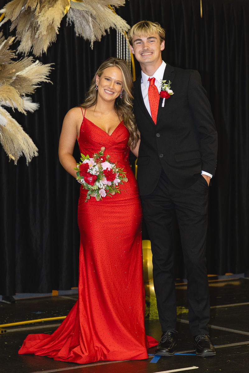 Couple in formal wear posing on stage: woman in red gown holding a bouquet, man in black suit with red tie and boutonniere, smiling.