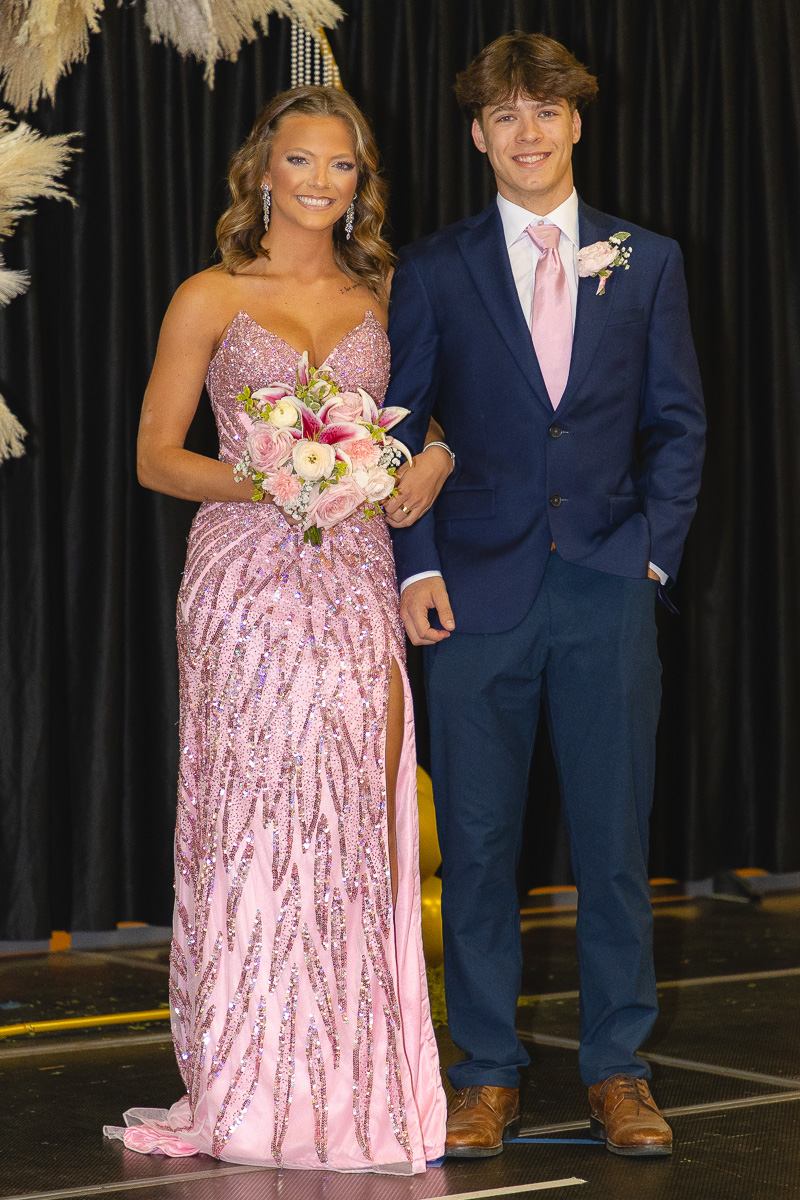 Young couple posing on stage in formal attire: woman in a pink sequin gown with a bouquet, man in a navy suit with a pink tie.