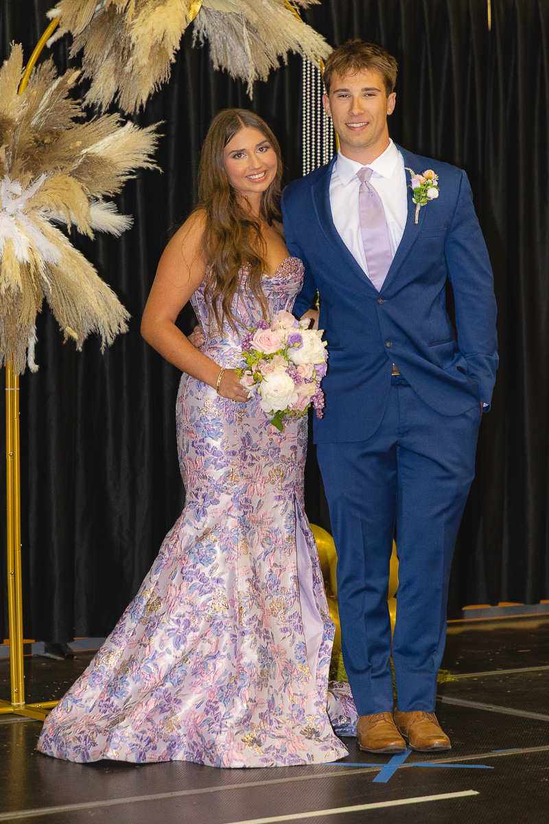 Couple posing in formal wear on stage: woman in a lavender floral gown holding a bouquet, man in a blue suit with a boutonniere. Prom couple posing for a photo on a dark backdrop with pampas grass decor.