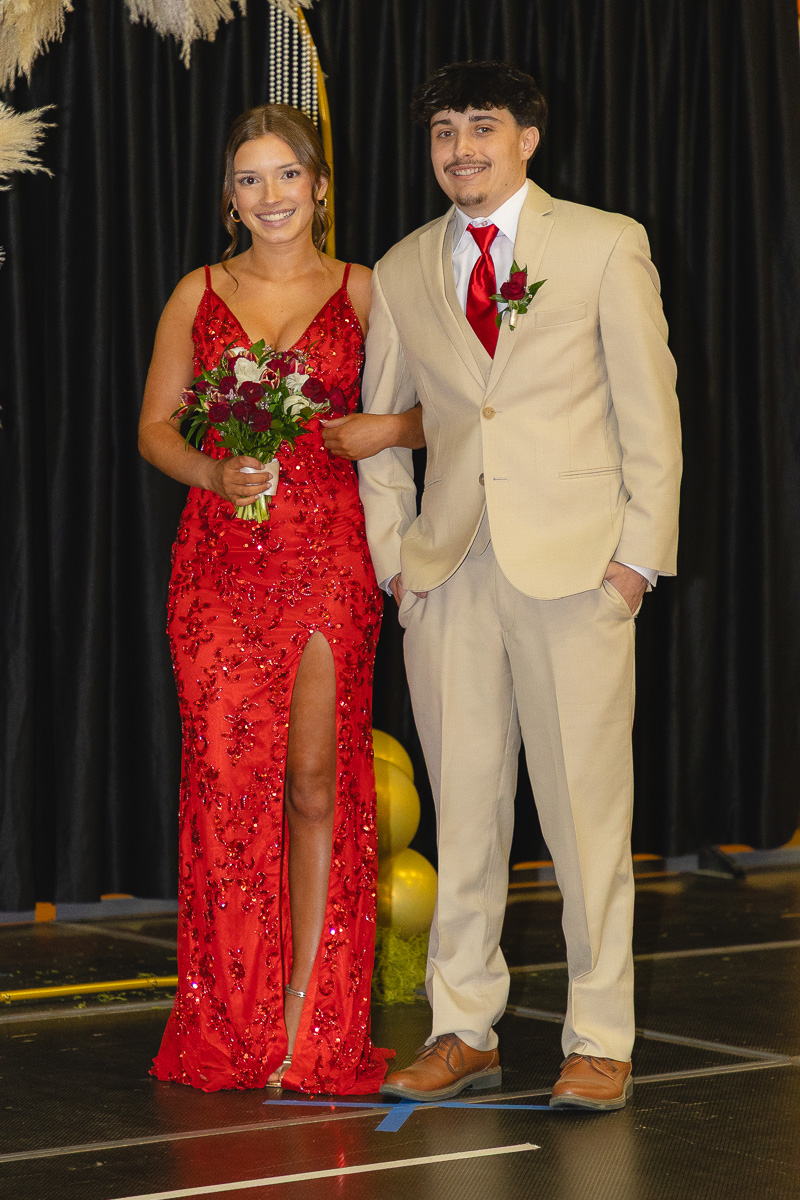 Couple posing on stage in formal wear: woman in red sequin gown with bouquet, man in beige suit with red tie and boutonniere, smiling.