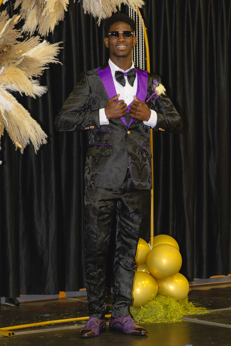 Man in a black patterned suit with purple lapels and bow tie posing on a stage with gold balloons behind him.