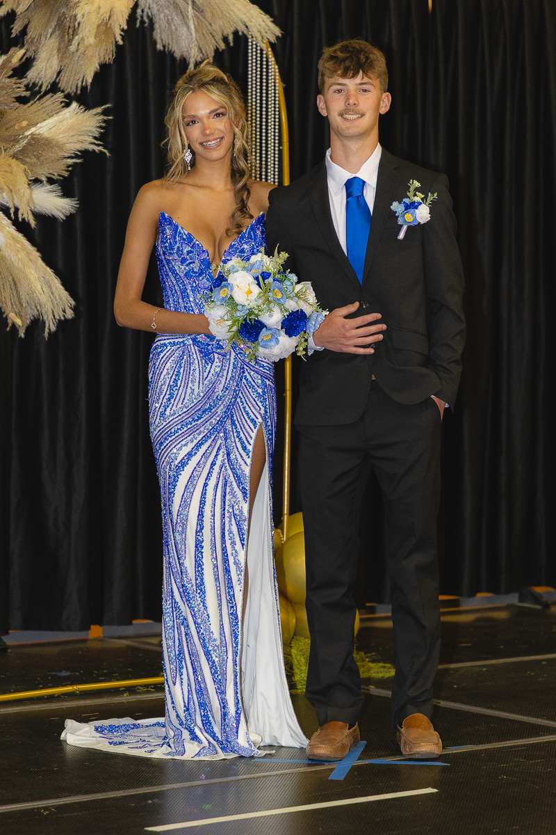 Young couple in formal wear posing on a stage; woman in a blue beaded gown holding a bouquet, man in a black suit with blue tie