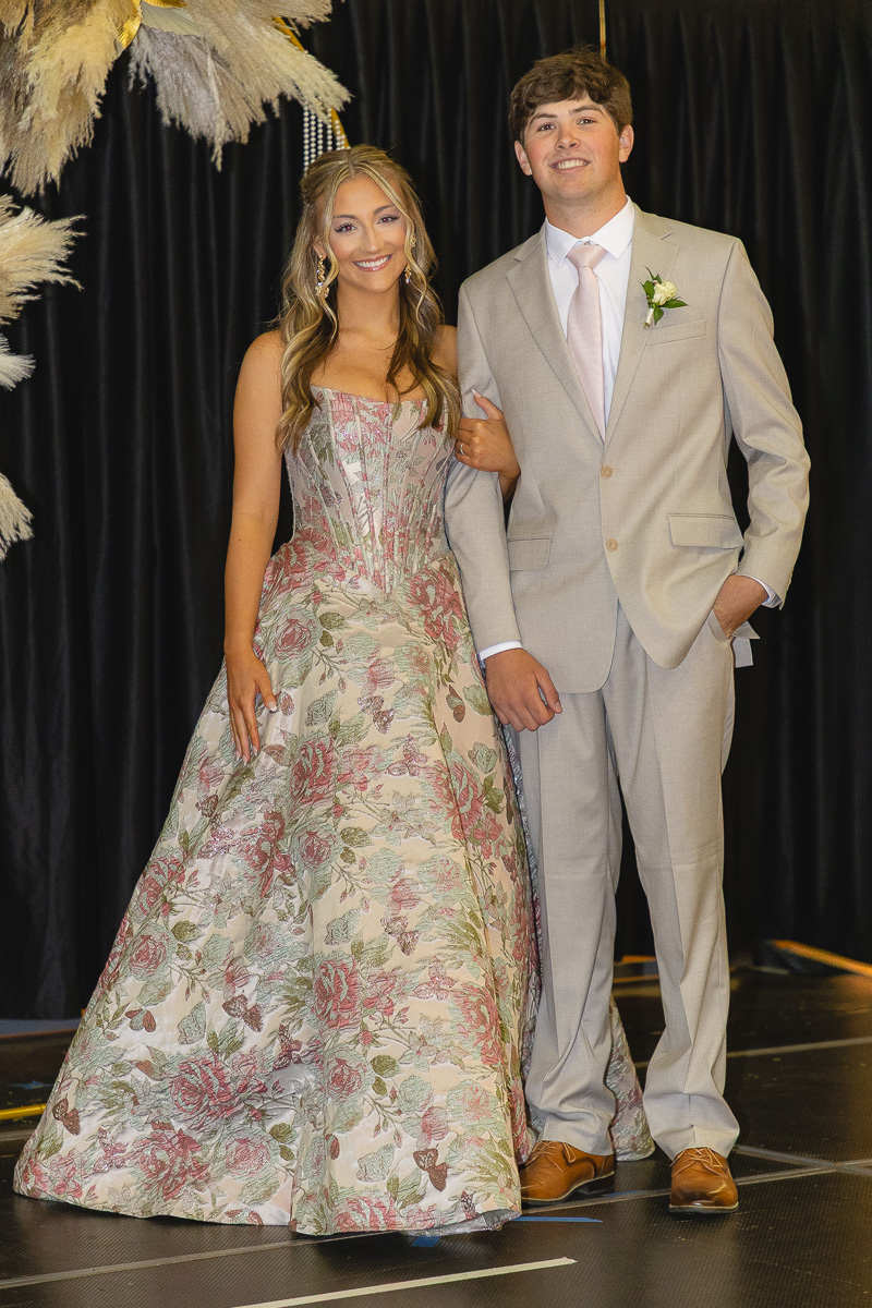 A smiling couple stands arm-in-arm on a stage: girl in a pink-floral ball gown and boy in a light gray suit with a pink tie and boutonniere.