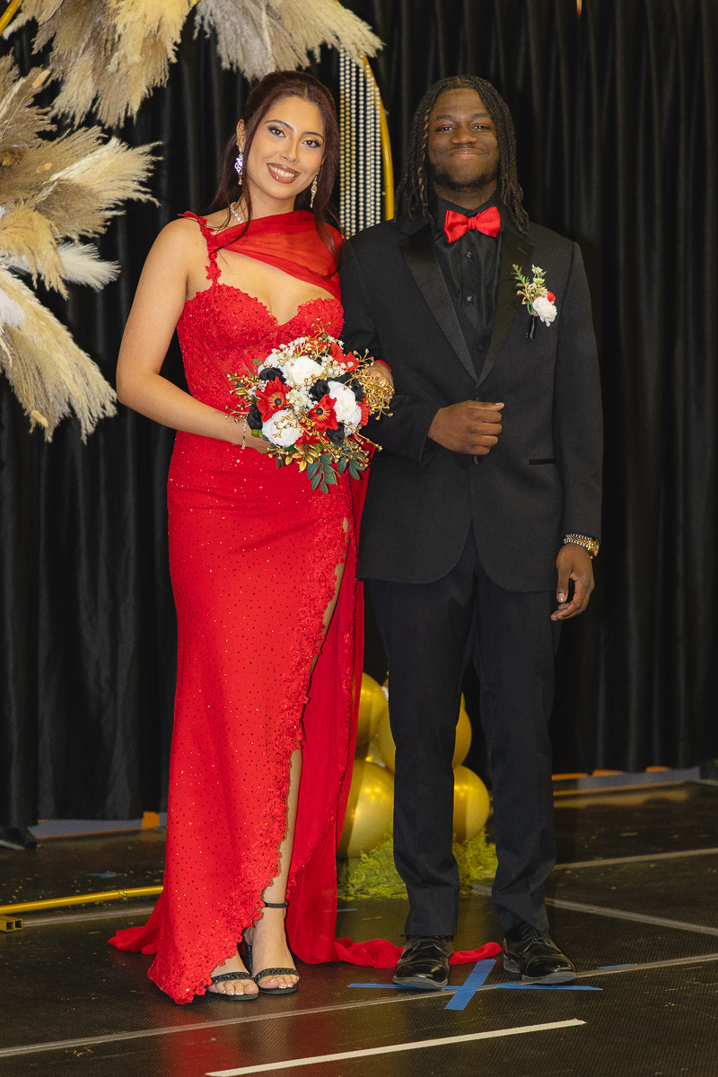 Couple posing on a stage: woman in a red sequined gown holding a bouquet, man in a black suit with a red bow tie and boutonniere.
