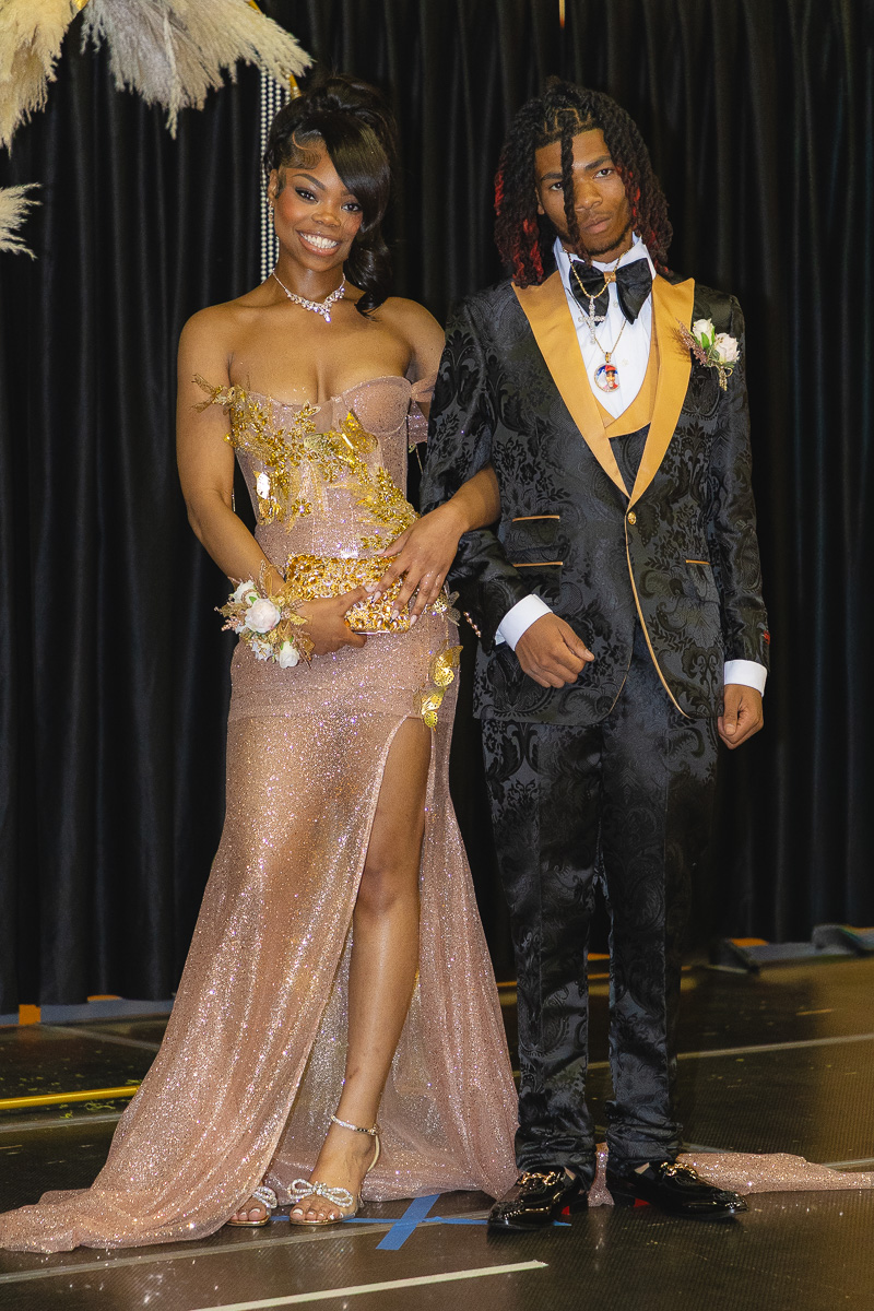 Young couple in formal wear posing on a stage; woman in a sparkling rose gown with slit, man in a black gold-trimmed suit and bow tie.