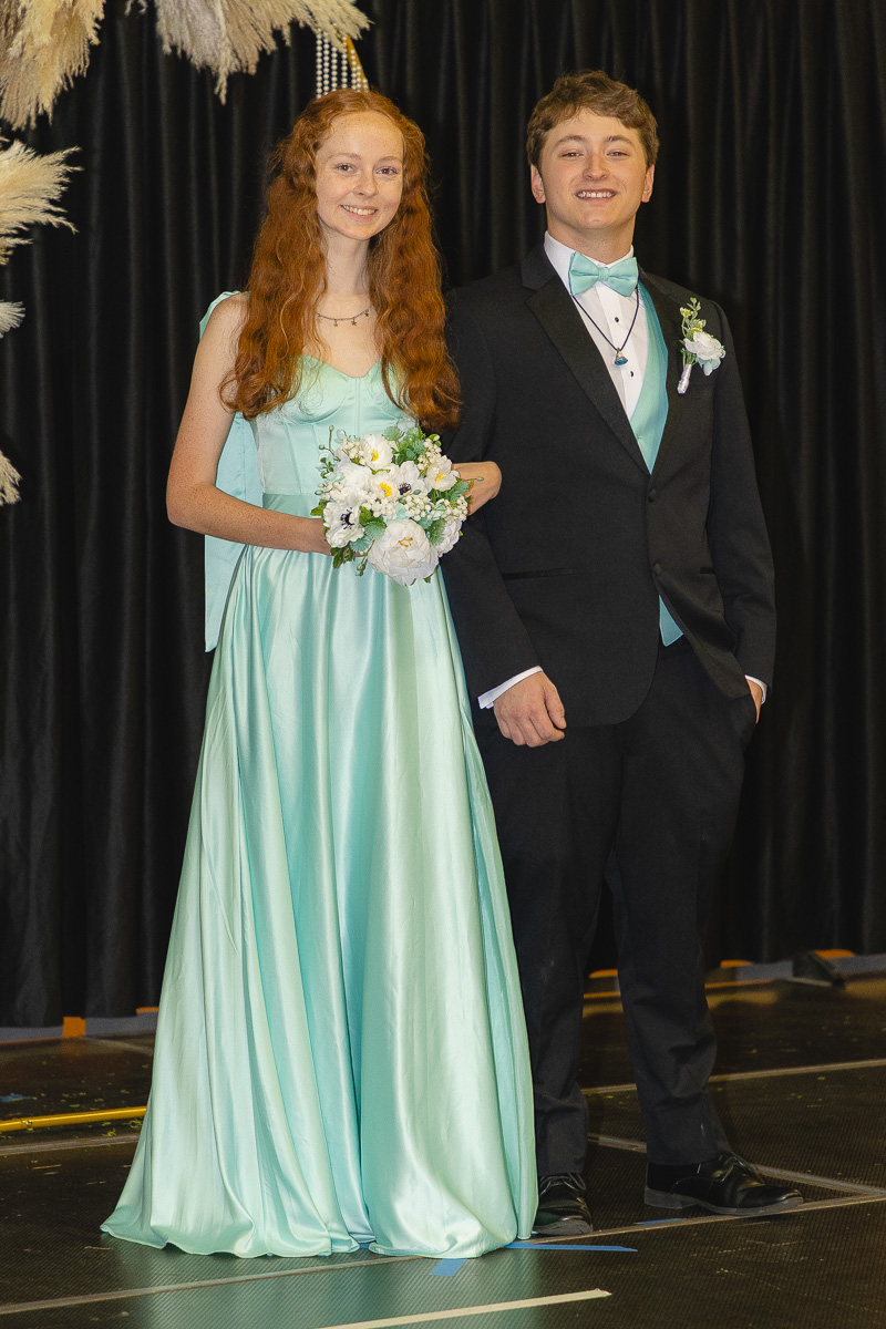 Two teens in formal attire pose on stage: a red-haired girl in a mint gown holding a white bouquet, beside a boy in a black suit with a turquoise bow tie.