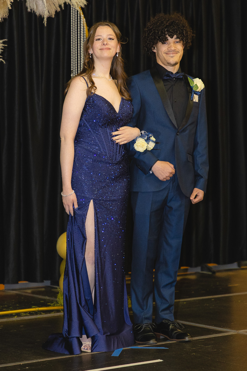 Two young adults in formal wear posing on a stage: a woman in a glittery blue gown with a thigh-high slit and a man in a blue suit with a boutonniere.
