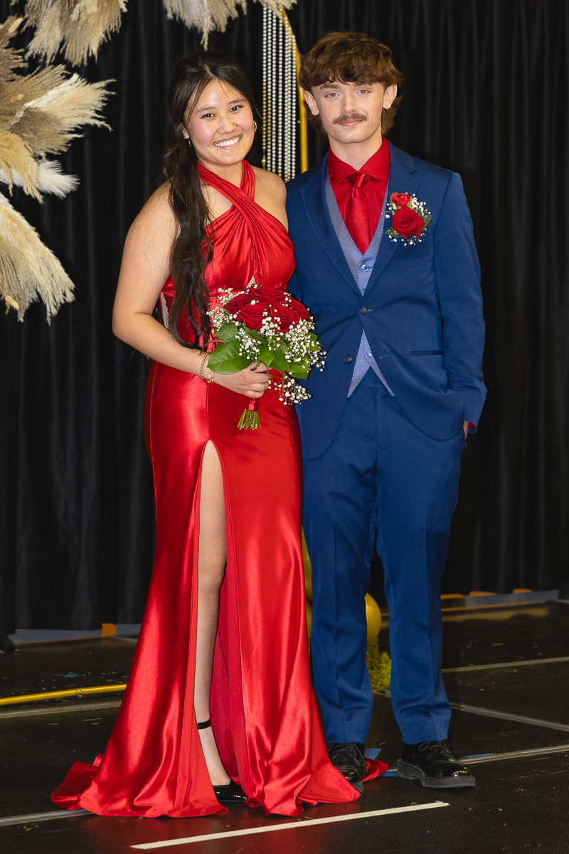 Smiling couple in formal attire posed on stage; woman in a red gown holding a bouquet of red roses, man in a blue suit with a red boutonniere.