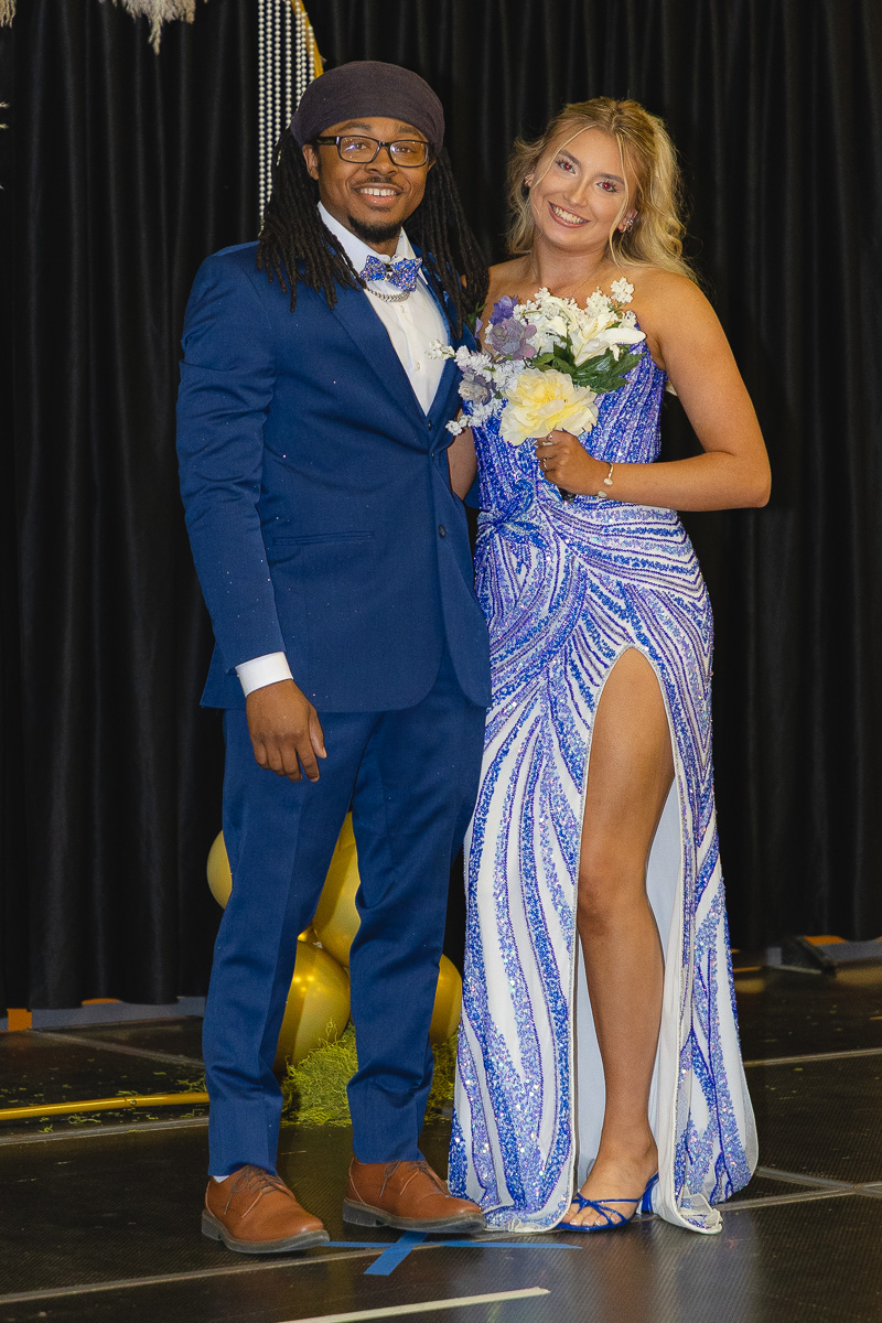 A smiling couple posing on stage: man in a blue suit and brown shoes, woman in a sparkling blue gown holding a bouquet.