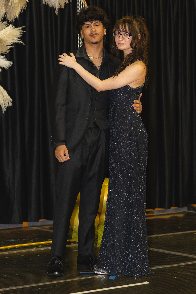 Young couple poses together on a stage, man in a black suit and woman in a sparkly navy gown, arms around each other.