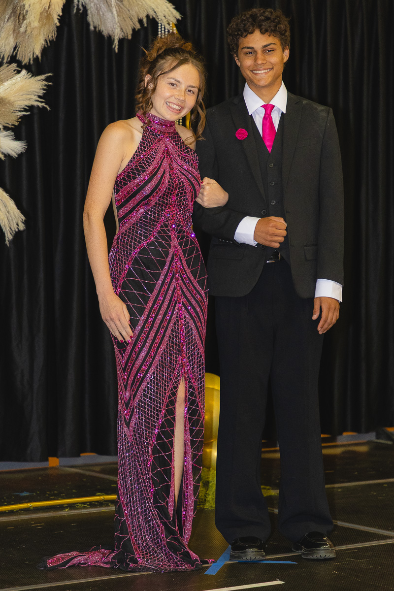 Teen girl in a sparkling pink gown stands arm-in-arm with a teen boy in a black suit on a stage with a dark curtain backdrop?