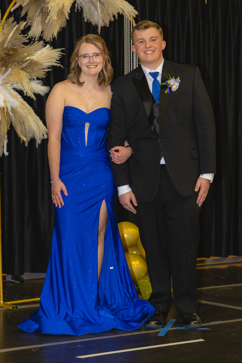 Couple posing in formal wear at a prom: woman in a blue strapless gown with slit and man in a black suit with blue tie on stage.