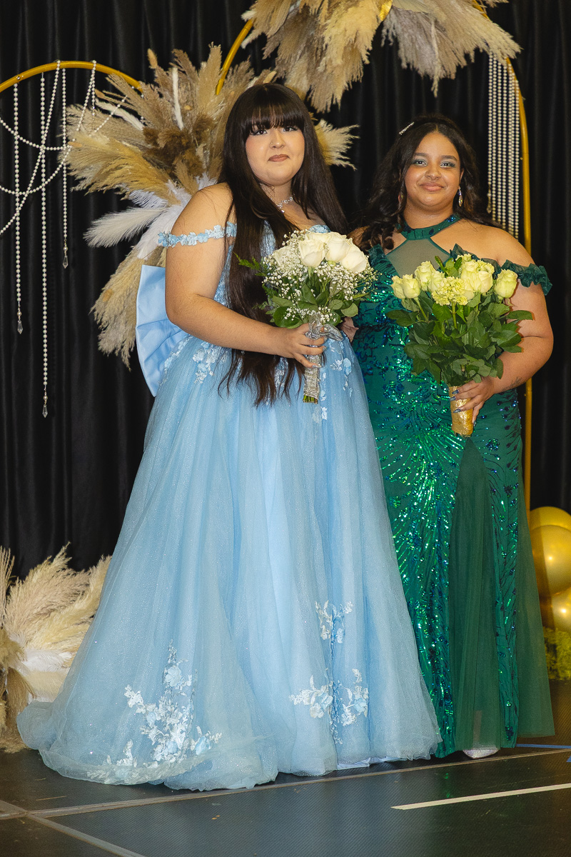 Two young women in formal gowns pose on a stage, each holding a bouquet of white roses.