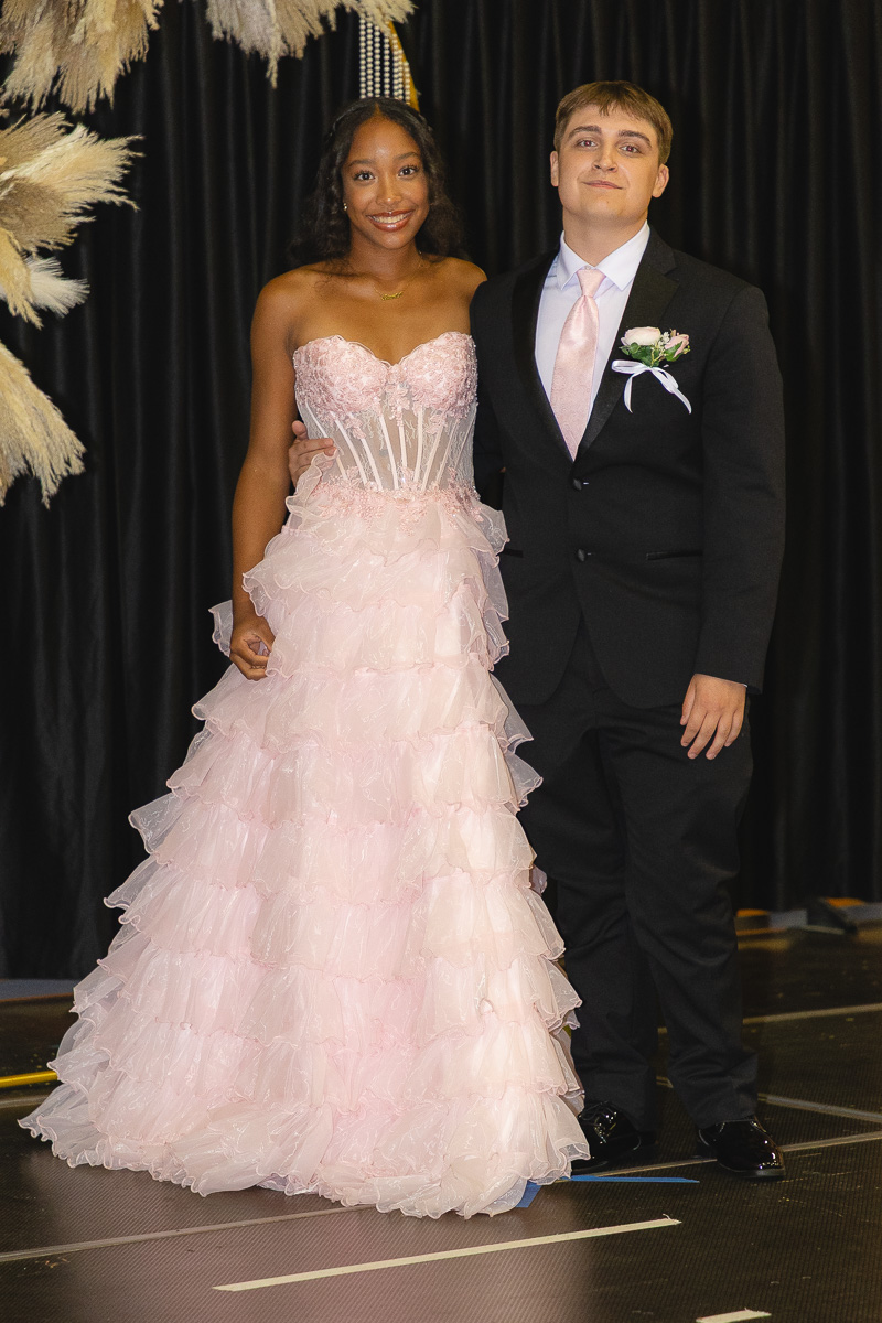 Teen couple posing in formal wear on a stage; girl in a pink strapless, ruffled ball gown and boy in a black suit with a pink tie and boutonniere.