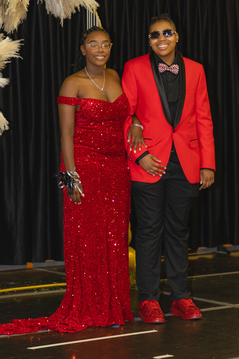 Two people in formal attire posing arm-in-arm on a stage: a woman in a red sequined off-shoulder gown and a man in a red blazer with a bow tie and sunglasses.