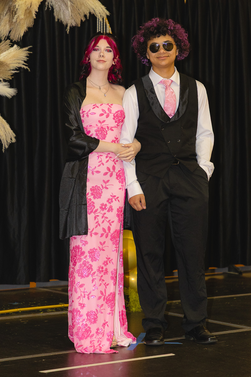 Two young people in formal wear pose arm-in-arm on a stage; the girl wears a pink floral gown with a black shawl and the boy wears a black vest and pants with a pink tie and sunglasses, against a dark backdrop with pampas grass.