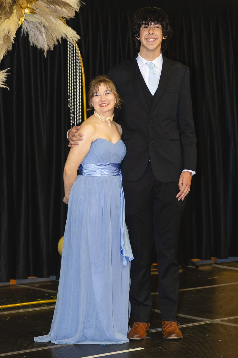 Two people in formal wear pose on a stage: a woman in a light blue gown and a man in a black suit with a light blue tie, smiling side by side.