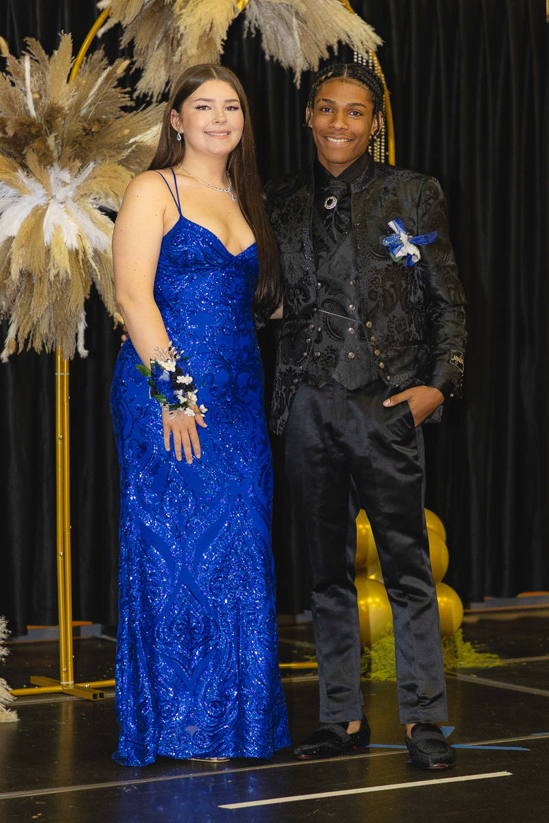 Two teens in formal attire posing together at a prom-like event: girl in a sparkling cobalt blue gown with a corsage, boy in a black patterned suit with boutonniere, smiling in front of a decorative backdrop.