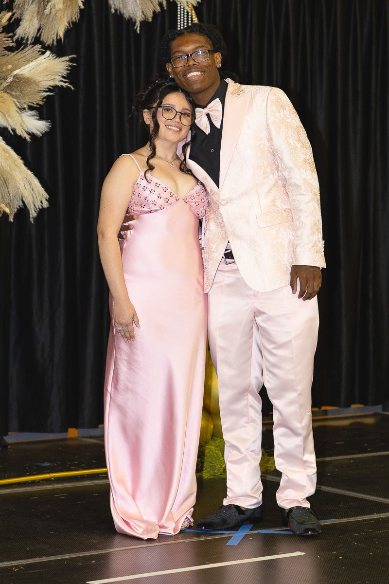 Two students in formal pink attire posing together on a stage; girl in a pink satin gown with glasses, boy in a pale pink suit and bow tie.