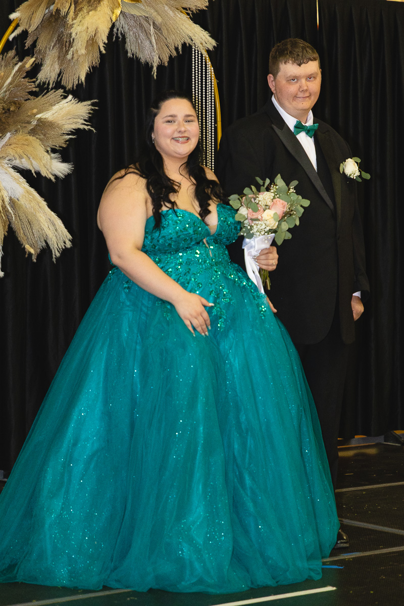 Couple posing at a formal event: woman in a teal glittery ball gown holding a bouquet, man in a black suit with a green bow tie.