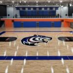 Empty indoor basketball court with a large tiger logo at center and blue bleachers in the background.